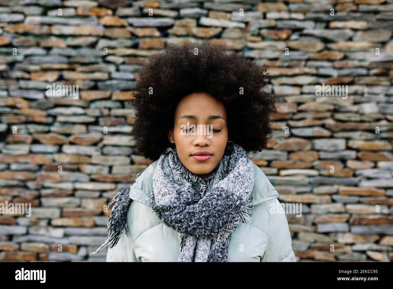 Afro junge Frau mit geschlossenen Augen gegen Steinmauer während Winter Stockfoto