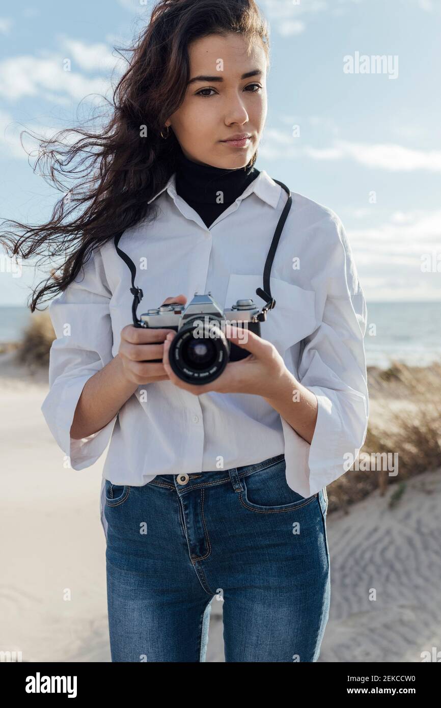 Schöne junge Frau, die Kamera hält, während sie am Strand steht Himmel Stockfoto Schöne junge Frau, die Kamera hält, während sie am Strand steht Himmel Stockfoto