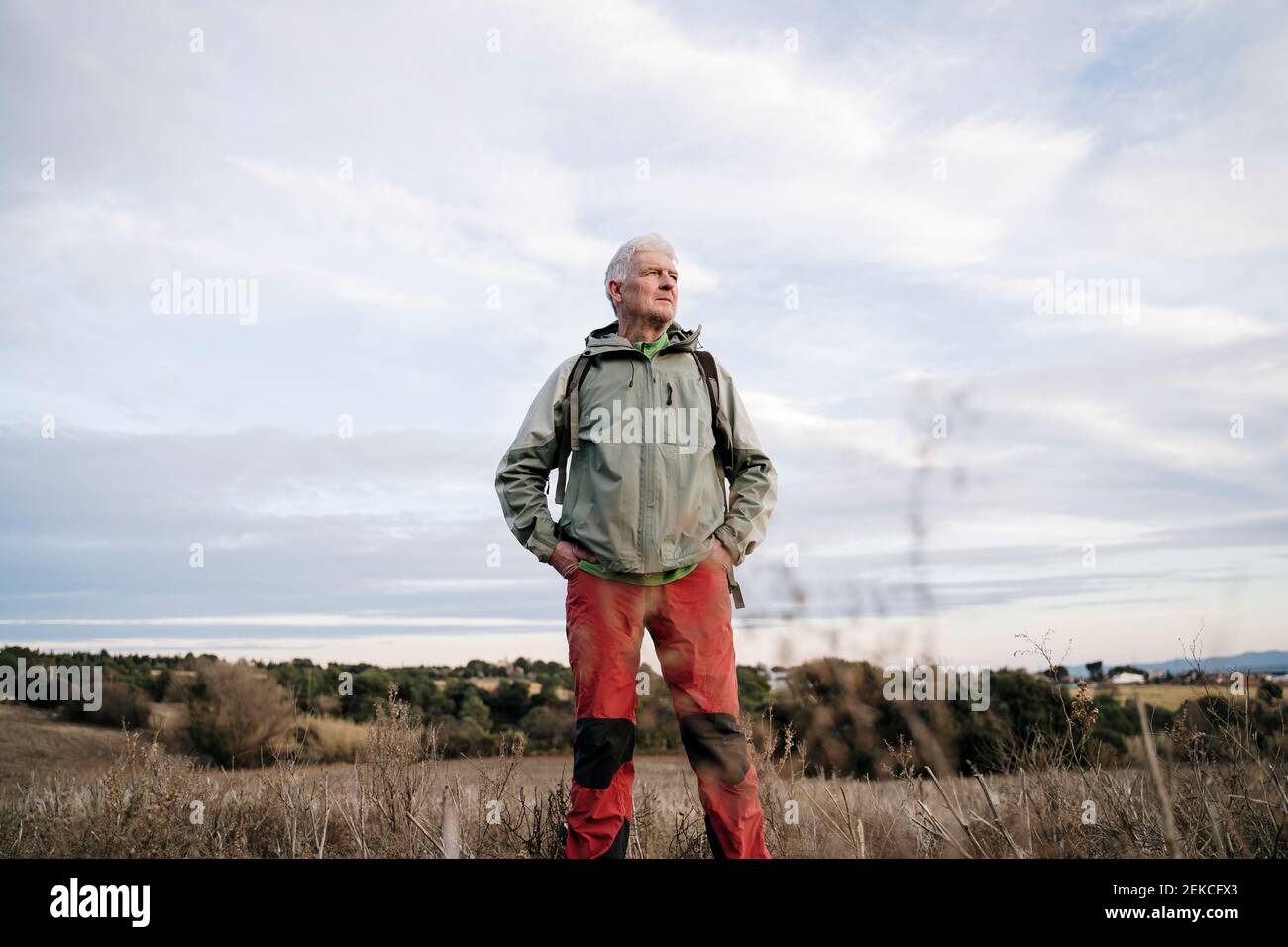 Älterer männlicher Wanderer mit Händen in den Taschen, die auf der Landwirtschaft stehen Feld gegen bewölkten Himmel auf dem Land Stockfoto
