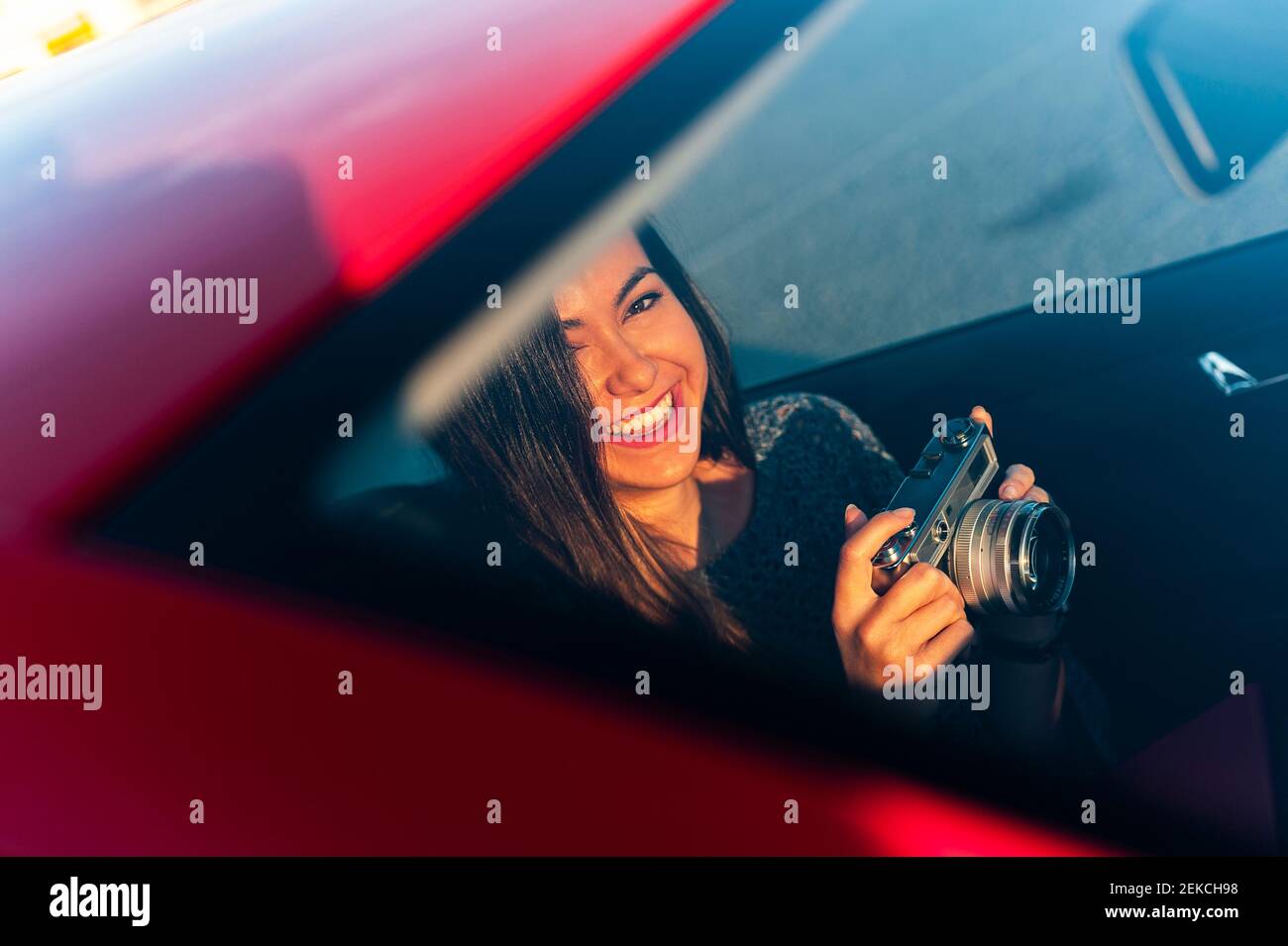 Lächelnde junge Frau mit Vintage-Kamera im Auto durch gesehen Windschutzscheibe an sonnigen Tagen Stockfoto Lächelnde junge Frau mit Vintage-Kamera im Auto durch gesehen Windschutzscheibe an sonnigen Tagen Stockfoto