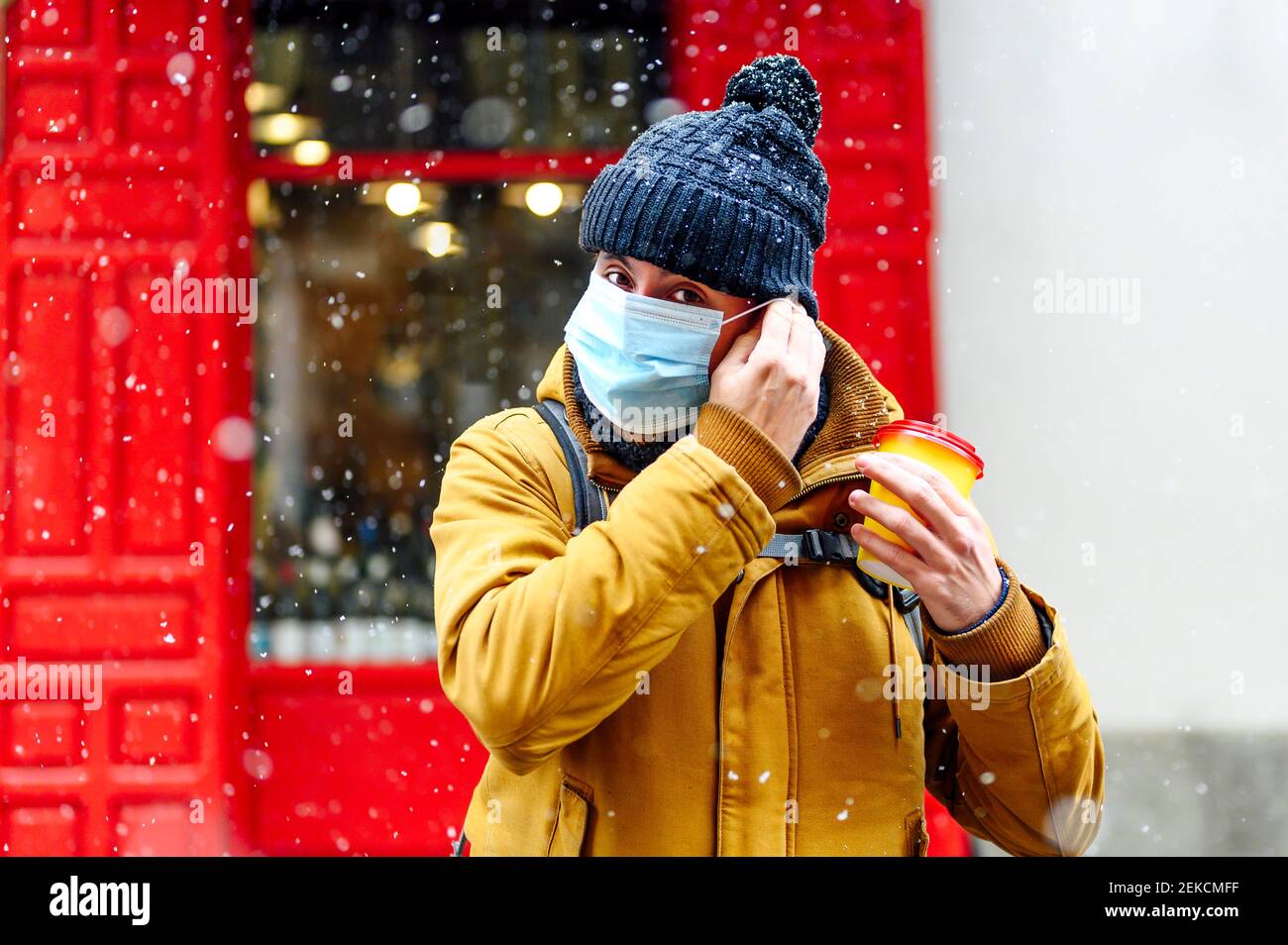 Mann in warmer Kleidung, der während des COVID-19 eine schützende Gesichtsmaske trägt, während er gegen den Laden steht Stockfoto