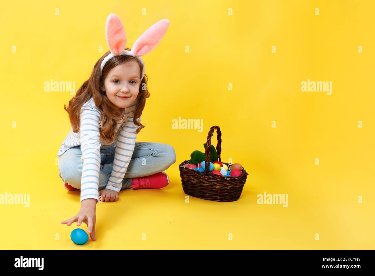 Kleines Mädchen in Hasenohren auf gelbem Hintergrund im Studio. Ein süßes Kind sitzt auf dem Boden mit einem Osterkorb und jagt nach einem Ei. Stockfoto