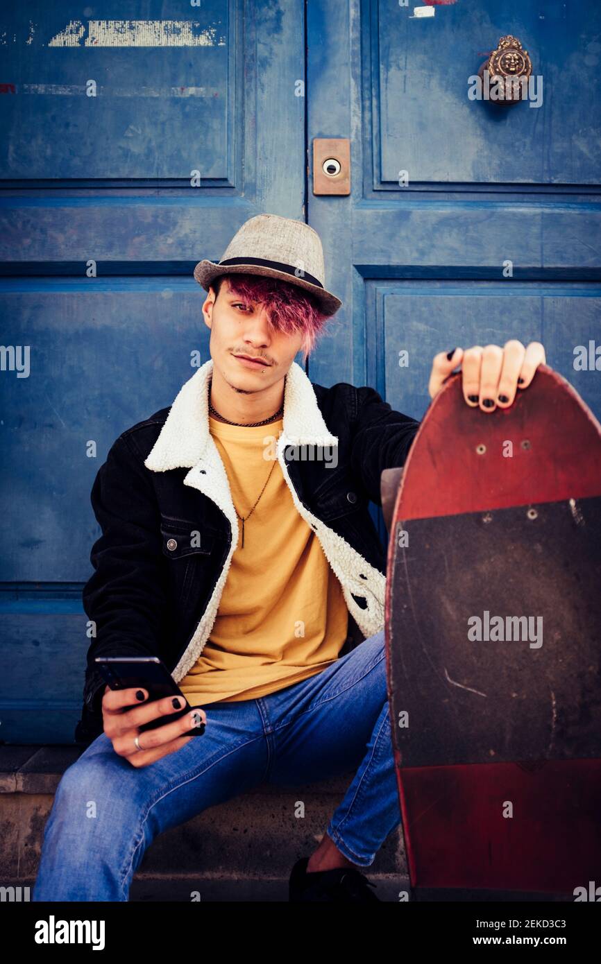 Diverse junge Mann mit violetten Haaren und alternative Look Stil Benutzen Sie ein modernes Telefon draußen mit blauen Türen im Hintergrund Und Skate Board, um mich zu bewegen Stockfoto