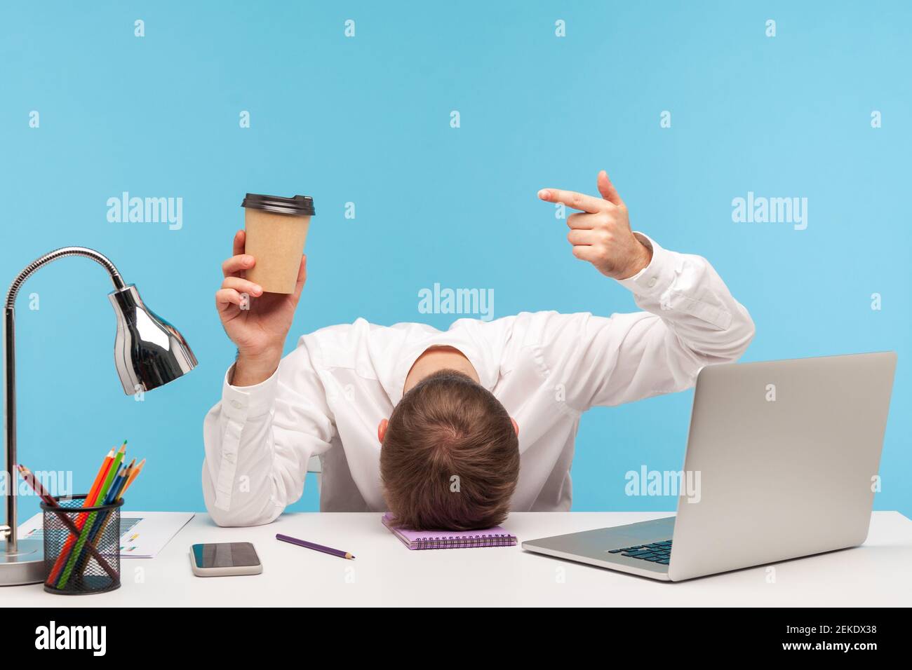 Ich brauche mehr Kaffee. Müder Geschäftsmann liegt mit dem Gesicht nach unten auf dem Tisch und zeigt auf Papierbecher in der Hand, Mangel an Energie, Überarbeit. Innenaufnahme im Studio Stockfoto