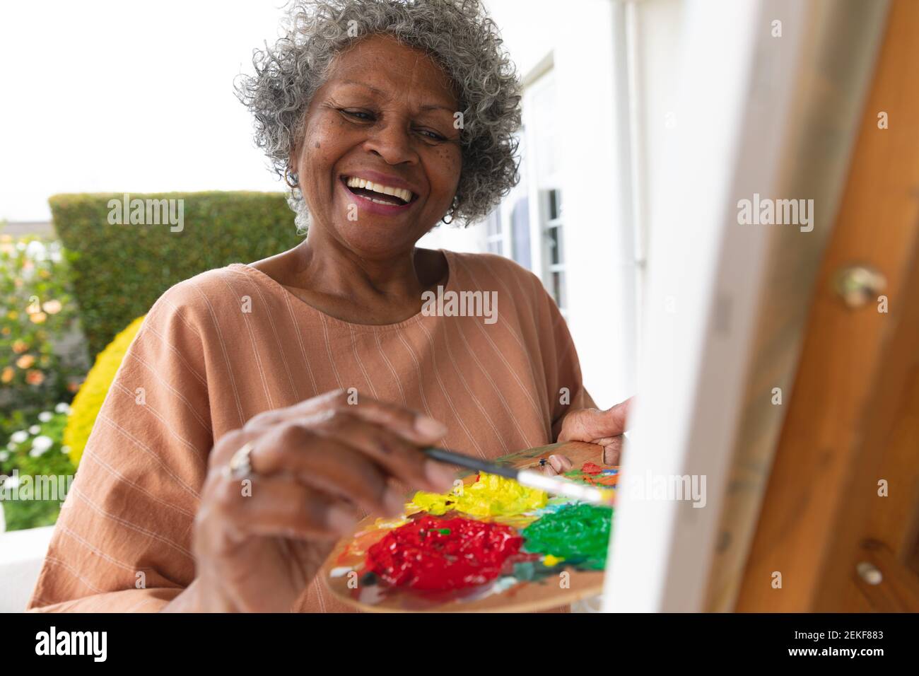 afroamerikanische ältere Frau lächelt beim Malen auf Leinwand stehend Auf der Veranda des Hauses Stockfoto