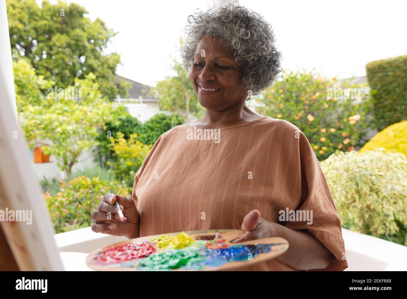afroamerikanische ältere Frau lächelt beim Malen auf Leinwand stehend Auf der Veranda des Hauses Stockfoto