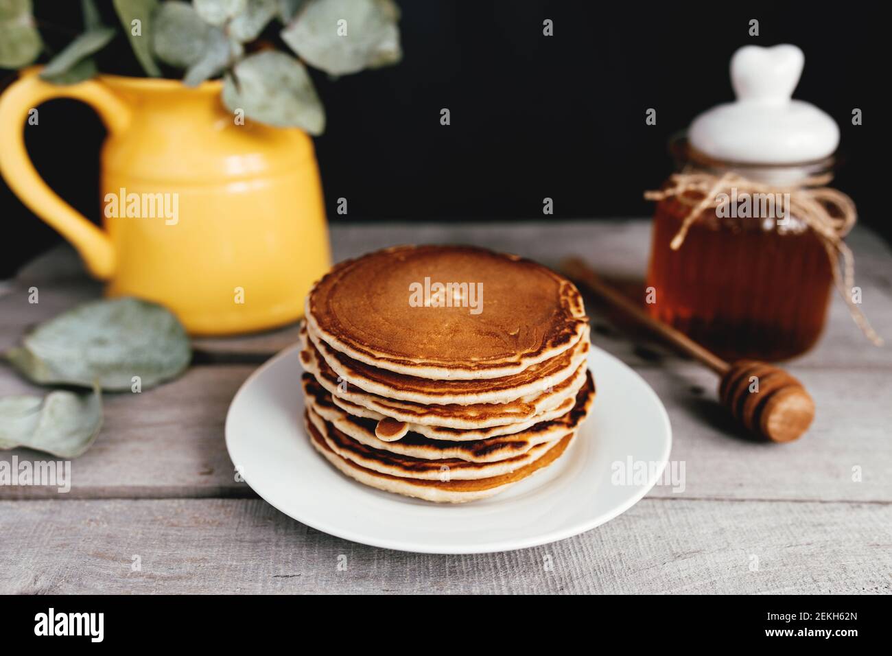 Saftige Pfannkuchen mit Honig auf einem weißen Teller, Löffel, Glas, Holztisch, gelbe Vase mit Eukalyptus. Hochwertige Fotos Stockfoto