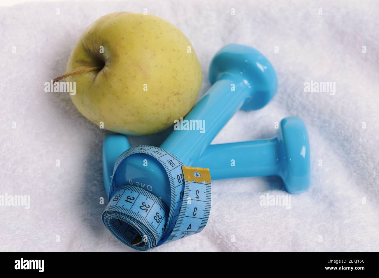 Gesundheits-Regime und Fitness-Symbole. Kurzhanteln nahe grünem Apfel auf weichem hellem Stoffhintergrund. Apfelfrucht und Langhanteln mit blauem Maßband gebunden. Gesundes Lifestyle- und Sportkonzept. Stockfoto