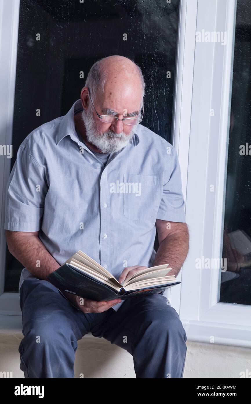 Älterer Mann, der nachts in seinem Wintergarten sitzt und einen Roman liest. Stockfoto