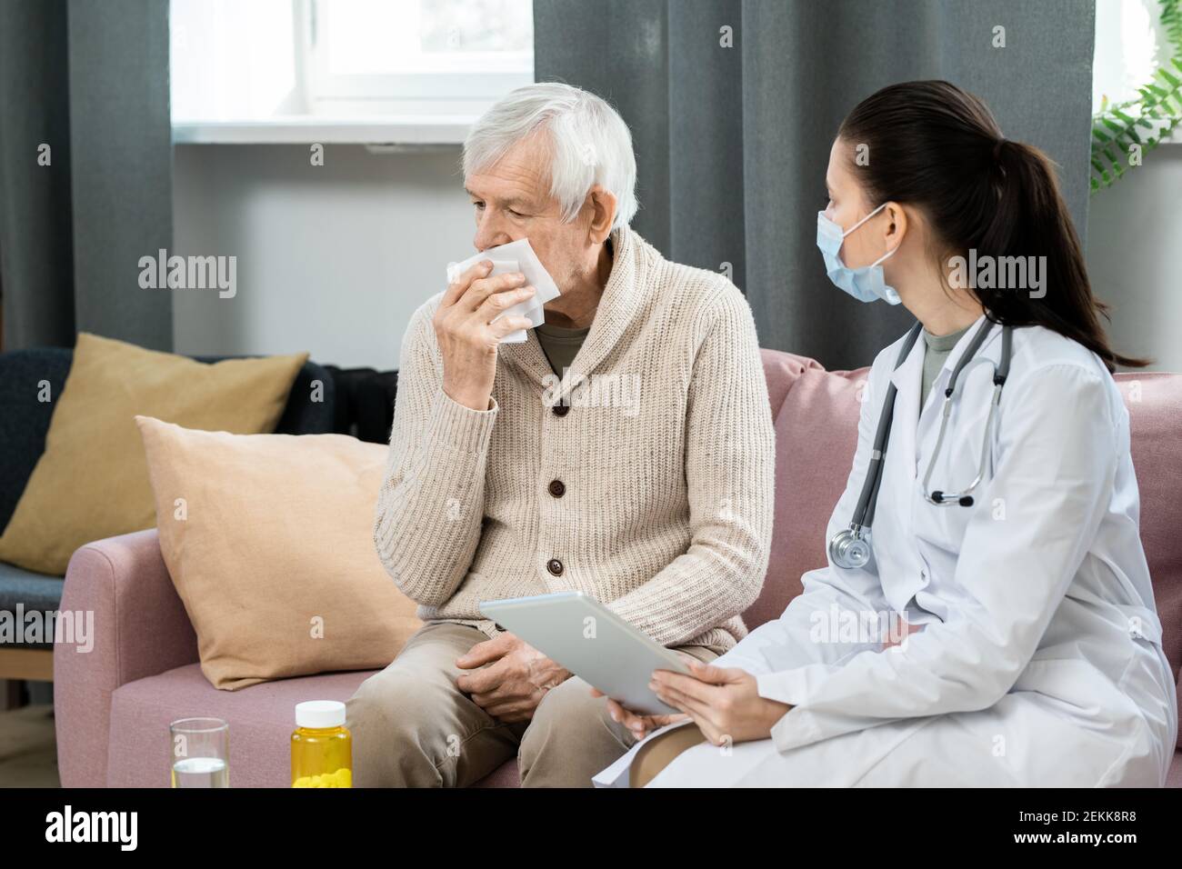 Junge Allgemeinärztin mit digitalem Tablet, die krank sieht Älterer Mann mit Taschentuch am Gesicht auf der Couch sitzend Vor ihr Stockfoto