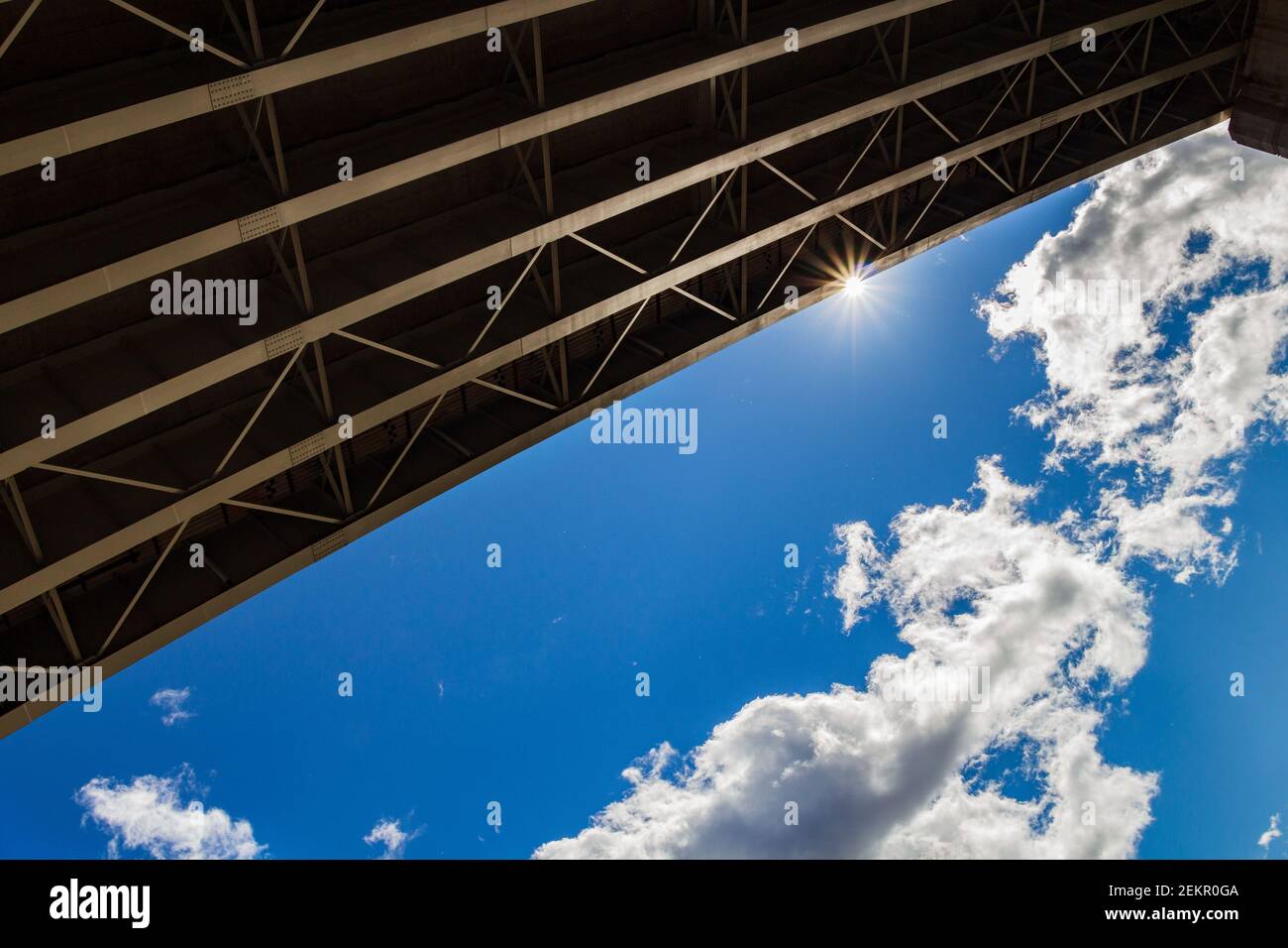 Die Linien von Stahlträgern unter der Überführung schaffen einen markanten Kontrast zum blauen Himmel, Wolken und Sonneneinstrahlung. Stockfoto