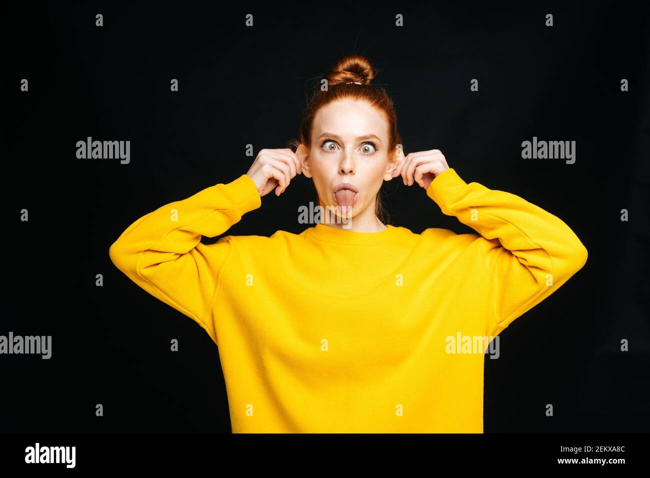 Verrückte fröhliche junge Frau in gelben Pullover berühren Ohren mit Hände und hervorstehende Zunge Stockfoto