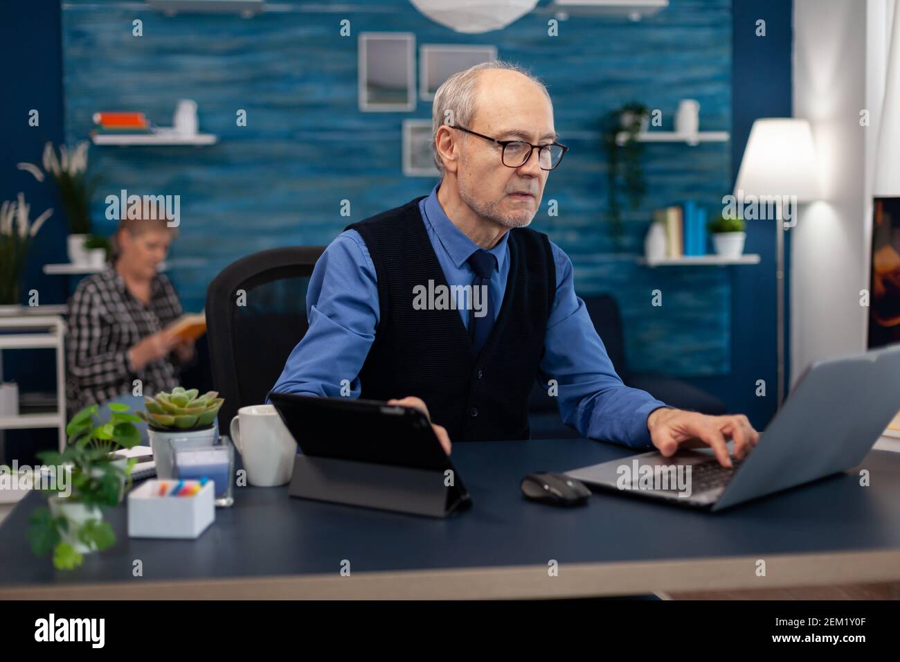 Leitender Manager, der an der Präsentation mit einem Laptop und Tablet-pc im Büro arbeitet. Ältere Mann Unternehmer in zu Hause Arbeitsplatz mit tragbaren Computer sitzen am Schreibtisch, während Frau liest ein Buch sitzen auf dem Sofa. Stockfoto