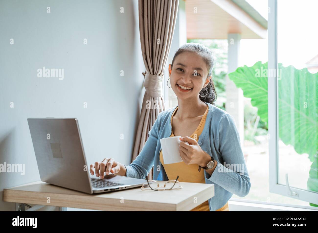 Selbstbewusste junge Frau in Smart Casual Wear arbeiten auf Laptop Während Sie zu Hause in der Nähe des Fensters sitzen Stockfoto