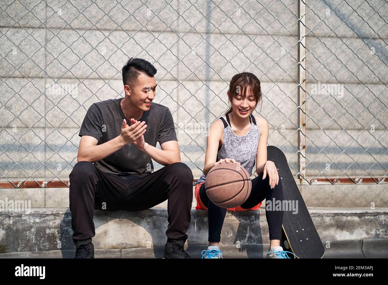 Junge asiatische Erwachsene männliche Basketballspieler und weibliche Skateboarder sitzen Auf der Gerichtsseite reden chatten Stockfoto
