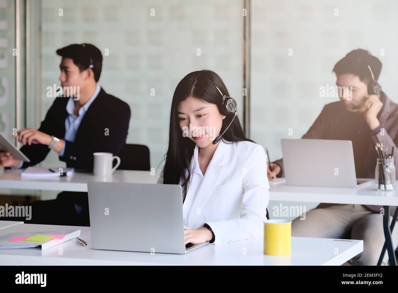Junge asiatische Frau, die im Callcenter arbeitet, umgeben von Kollegen. Stockfoto