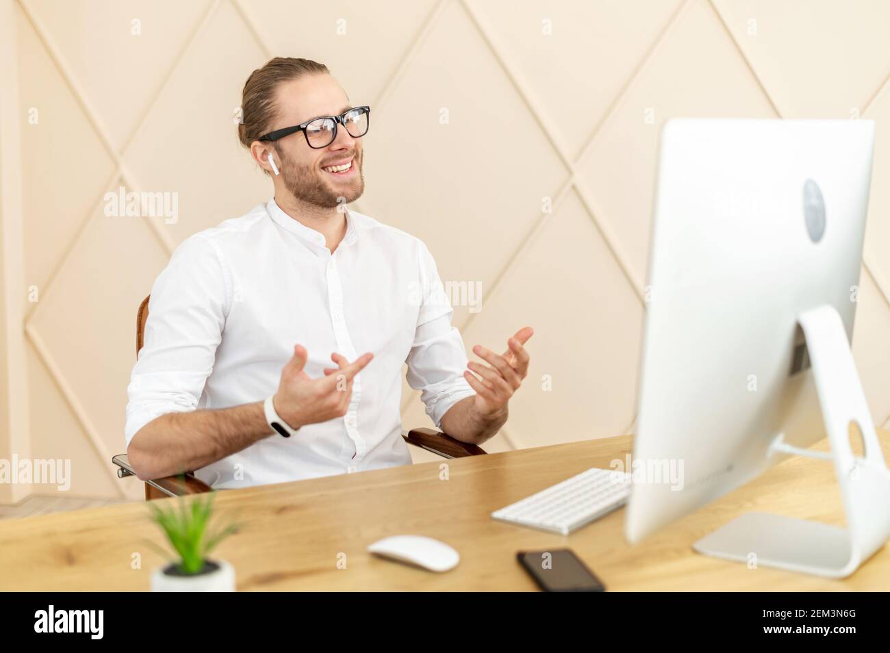 Selbstbewusster Mann mit hellbraunem Haar und Brille in weißem Hemd, lächelt aufrichtig, spricht am Computer, diskutiert Arbeitsprozess, Distanz Stockfoto