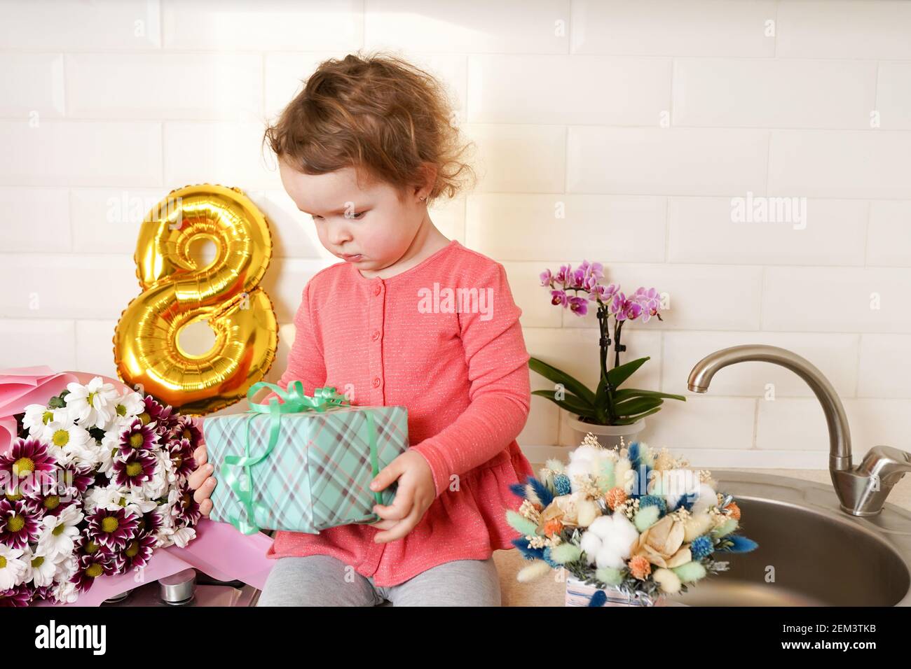 Nette lustige kleine Mädchen halten Geschenk-Box sitzen auf Küchentisch mit Blumensträußen Frühling Blumen und goldenen Ballon 8 acht , Gruß Mutter oder Großmutter Stockfoto