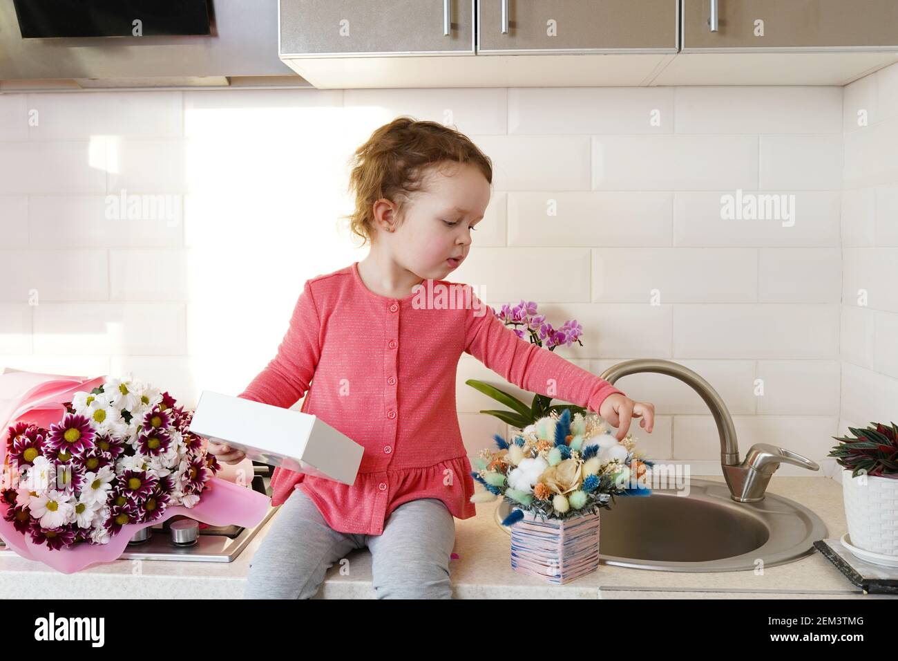 Nette lustige kleine Mädchen halten Geschenk-Box sitzen auf Küchentisch mit Blumensträußen Frühling Blumen und goldenen Ballon 8 acht , Gruß Mutter oder Großmutter Stockfoto