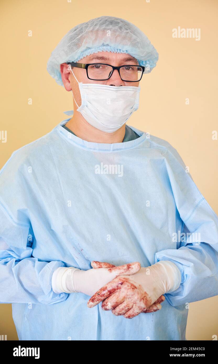 Porträt eines männlichen Arztes in einer Brille, der die Kamera anschaut. Mann Chirurg trägt sterile Handschuhe, blaue OP-Uniform, medizinische Kappe, Schutzmaske Gesicht. Konzept der Medizin, des Gesundheitswesens und der Chirurgie. Stockfoto
