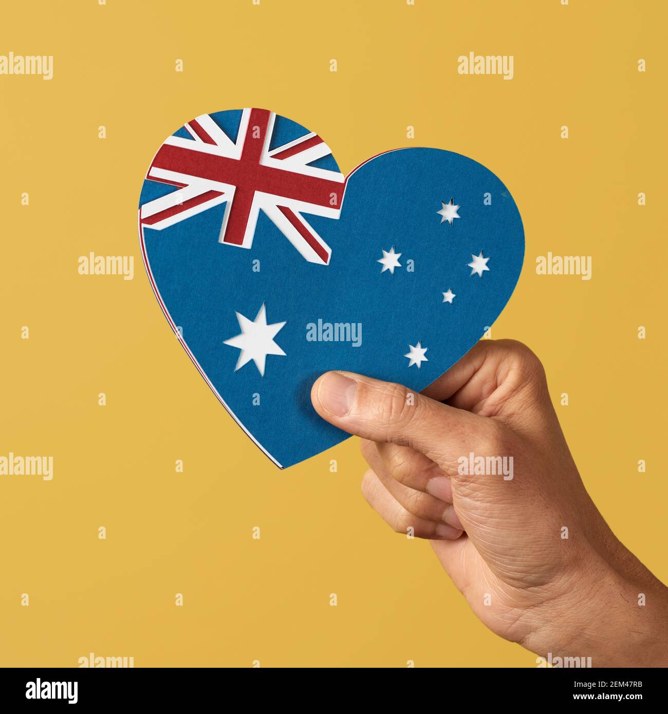 Nahaufnahme der Hand eines Mannes, der eine australische Flagge in Form eines Herzens hält, mit Ausschnitten aus Papier verschiedener Farben, auf einem gelben Hintergrund Stockfoto