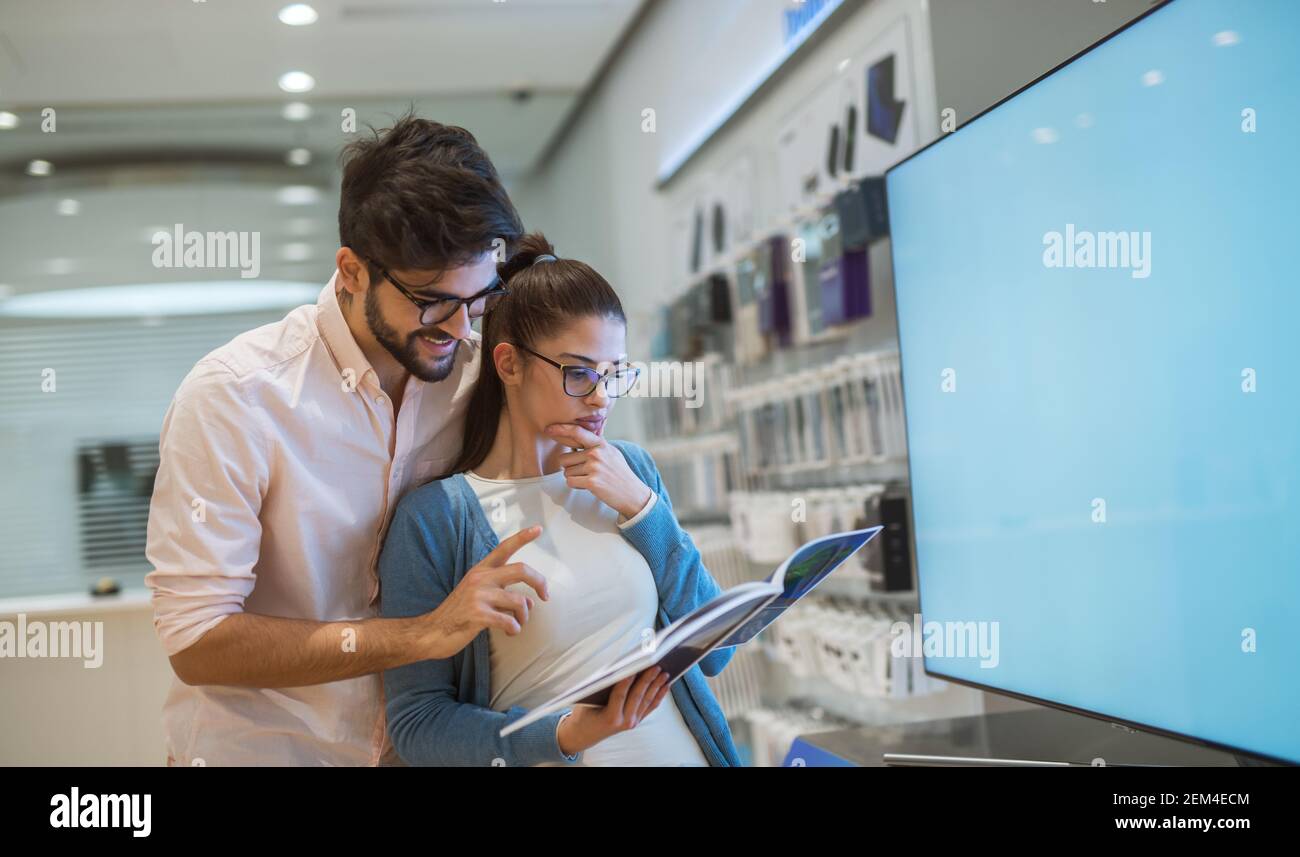 Nahaufnahme von attraktiven glücklich aufgeregt Hipster junge Liebe Das Paar stand vor dem großen Smart tv Während Mädchen halten Spezifikation in Stockfoto