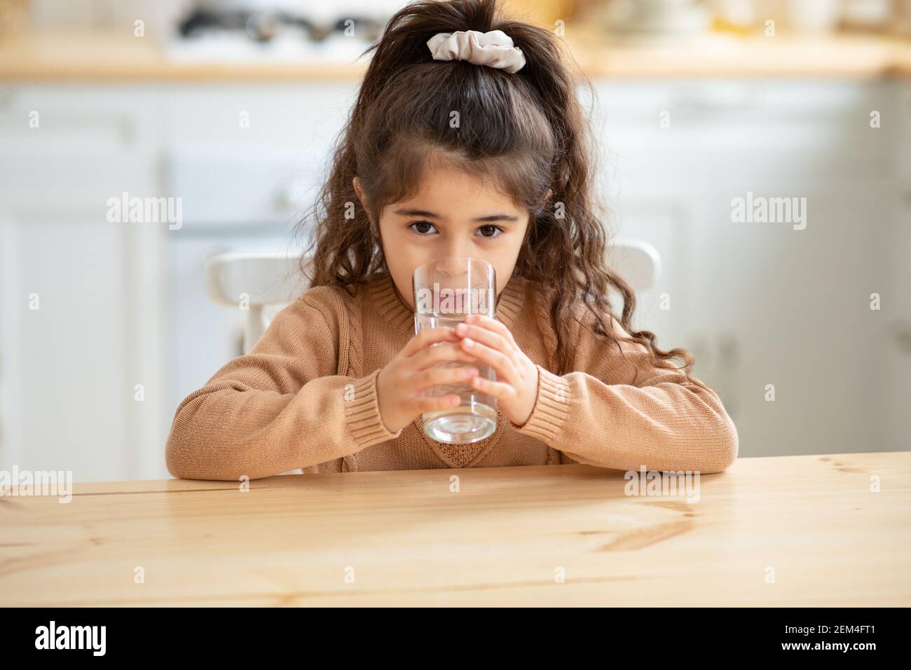 Nettes Kleines Mädchen, Das Wasser Aus Glas Trinkt, Am Tisch In Der Küche Sitzend Stockfoto