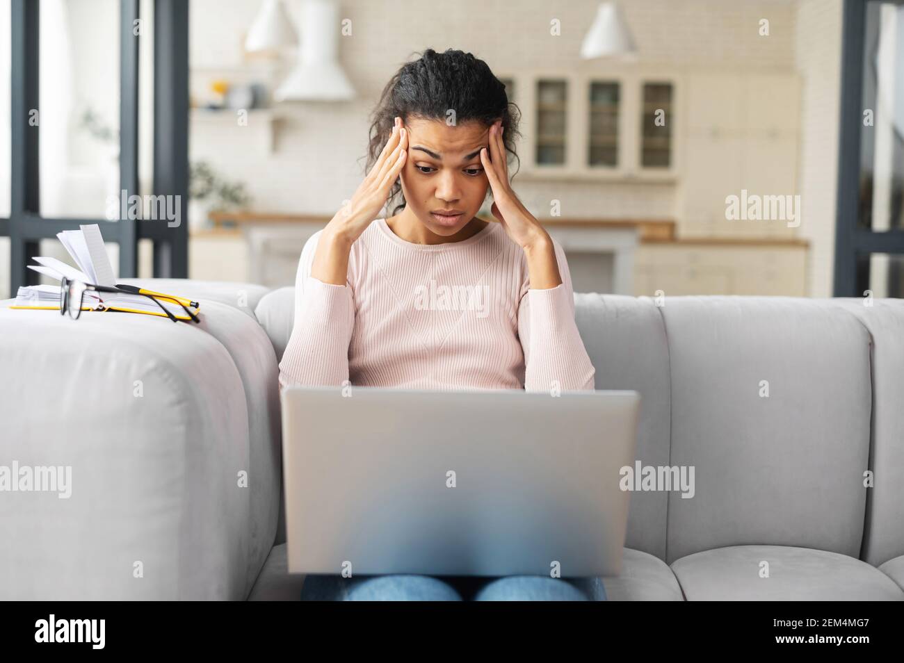 Mixed-Race-Freiberuflerin oder Studentin mit lockigen Haaren auf der Couch sitzend, müde von der ständigen Arbeit am Laptop, entfernt ihre Brille, berühren Tempel, Kopfschmerzen und kann sich nicht konzentrieren Stockfoto