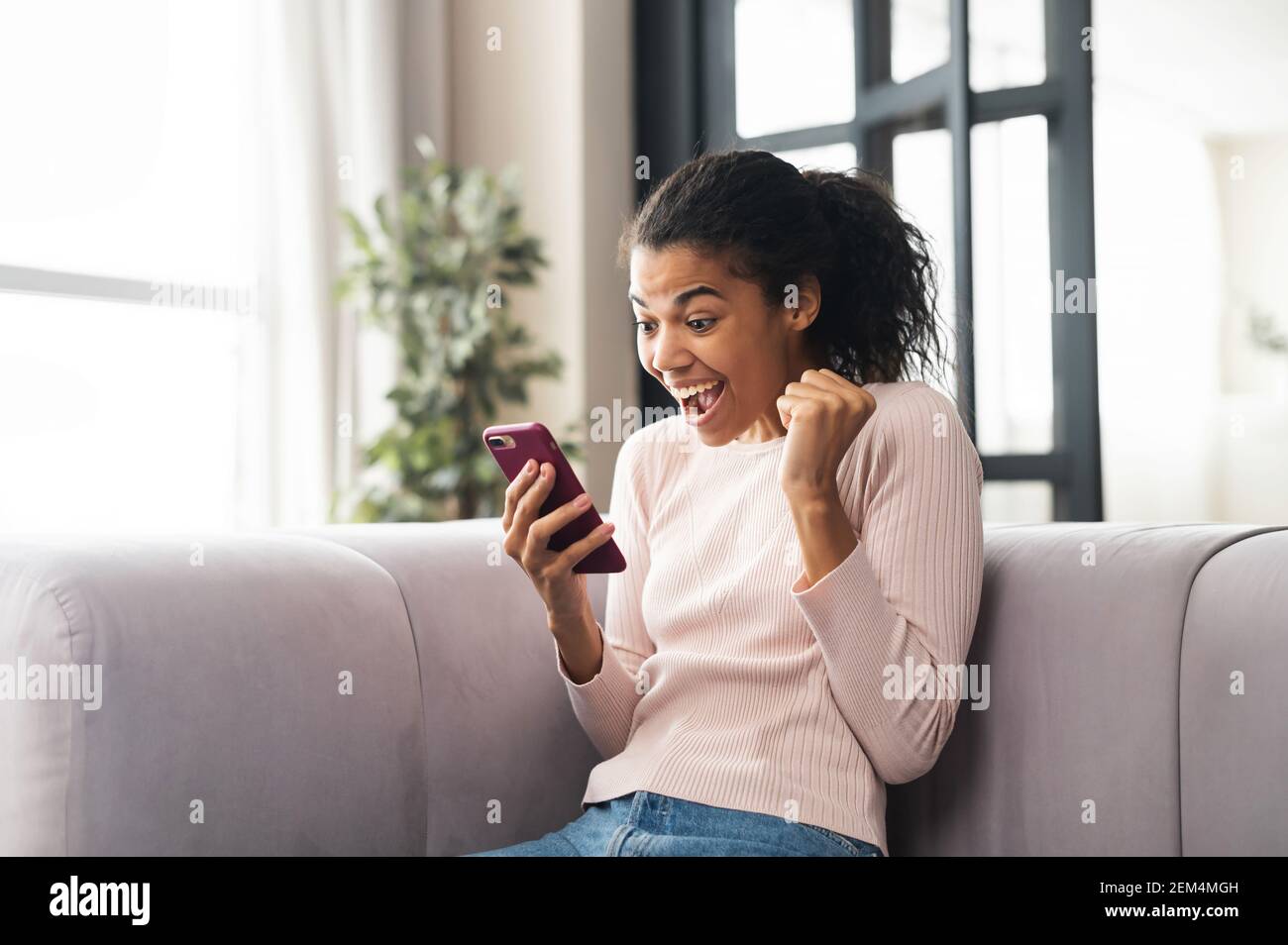 Aufgeregt Mixed-Rennen Teenager-Mädchen feiern Erfolg, Gewinner des Wettbewerbs zeigen Hand Faust und Blick auf Handy, sitzen auf der Couch und schreien ja, bestanden den Test, Ziel erreicht Stockfoto