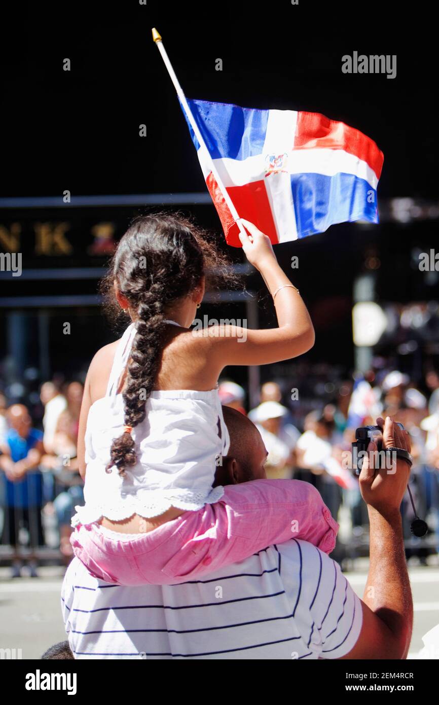Rückansicht eines Mädchens, das auf den Schultern ihres Vaters sitzt Und hält eine Flagge Stockfoto