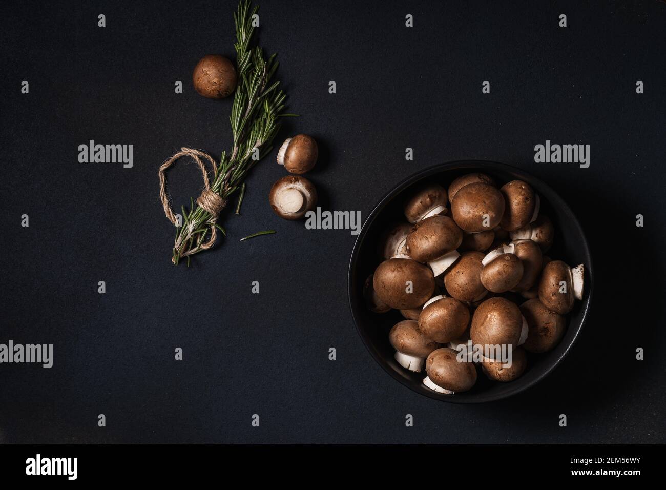 Schüssel mit rohen Baby bella Pilze und Bund von frischem Rosmarin auf dunkelgrauem Hintergrund von oben gesehen, pflanzliche Nahrung Stockfoto