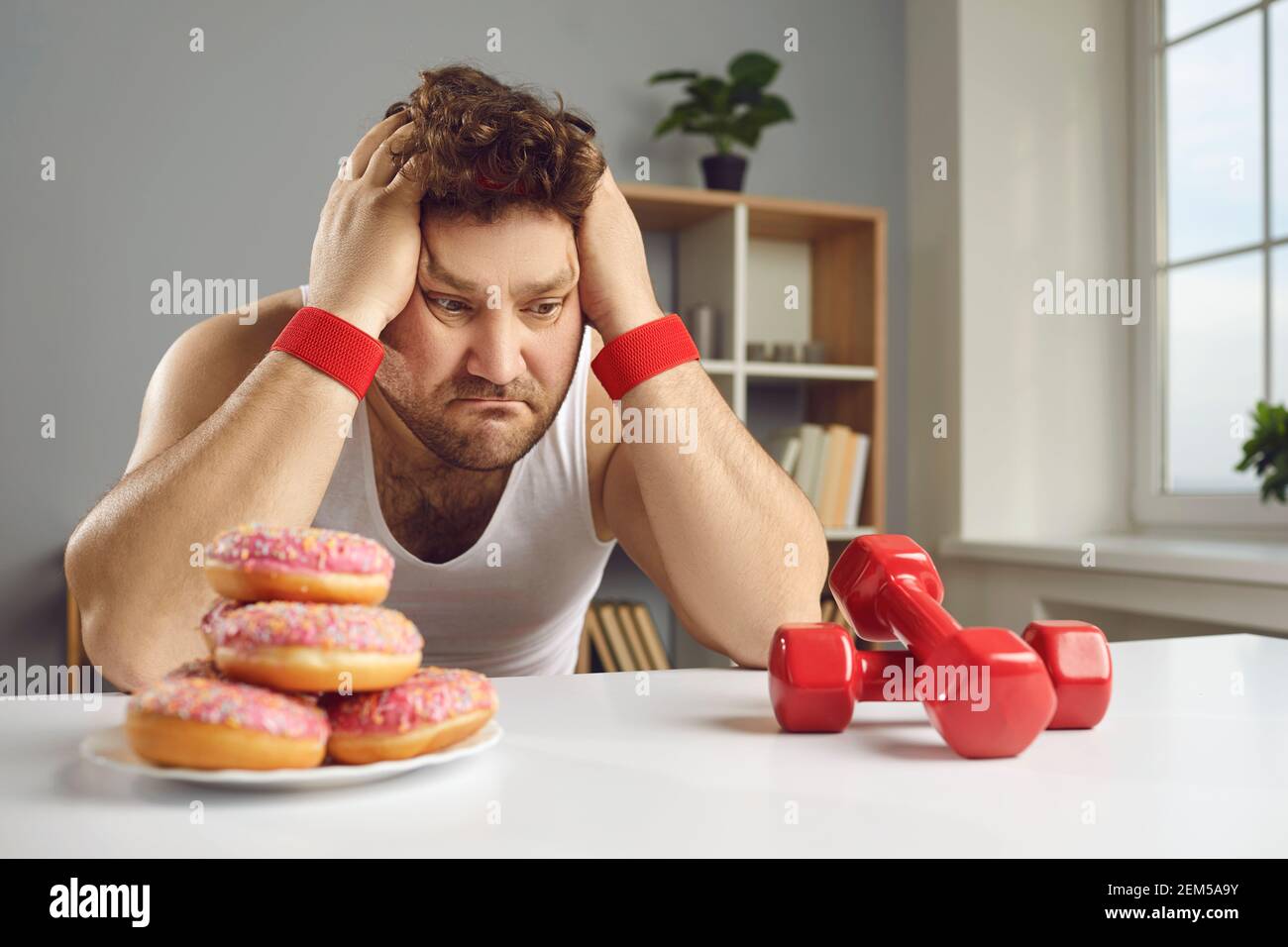 Trauriger Mann mit Blick auf Hanteln und Donuts Wahl zwischen gesund Und ungesunder Lebensstil Stockfoto