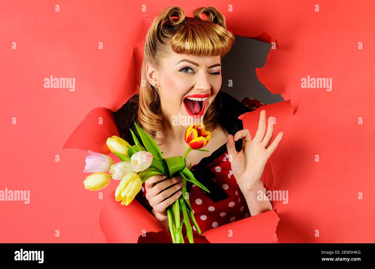 Winkende Frau mit Tulpen zeigt Schild ok. Lächelnd Pinup Mädchen durch Papier suchen. Glückliches Weibchen mit Blumenstrauß. Stockfoto