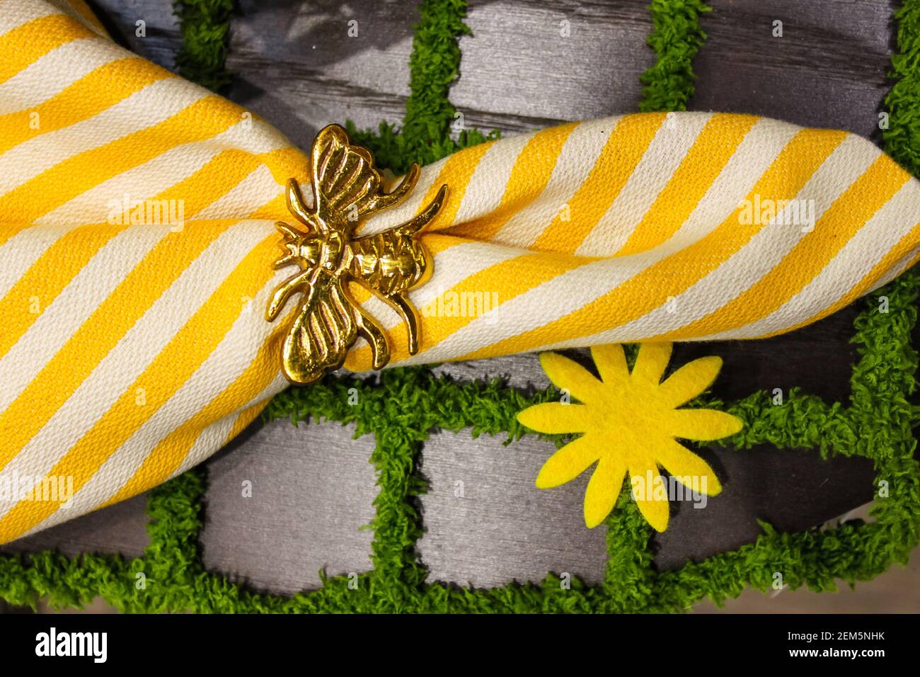 Gelb-weiße, knackige Leinenserviette mit goldener Bienenserviette Ring auf einem Tischset, das aussieht wie Pflastersteine mit Gras – Frühjahrsthema Stockfoto