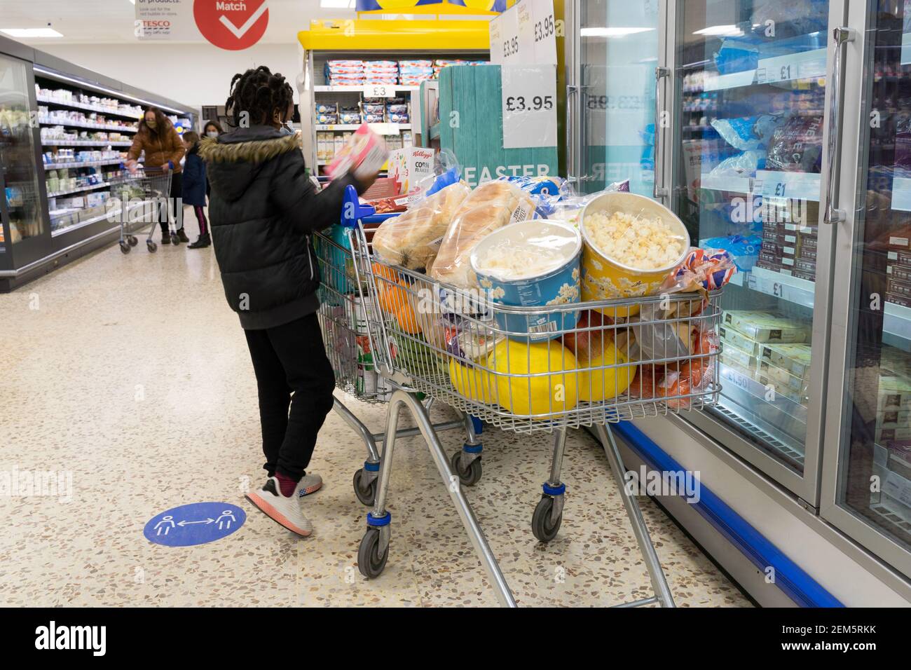 Ein Shopper legt mehr Artikel auf zwei Einkaufswagen voller Lebensmittel, Brot, Melonen und Popcorn, im Tesco Supermarkt, Großbritannien Stockfoto