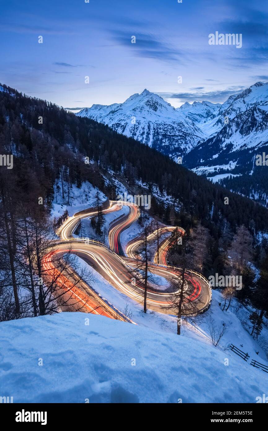 Autos leichte Wege auf den kurvenreichen Kurven der Malojapassstraße im Winter bei Sonnenuntergang, Bregaglia Tal, Kanton Graubünden, Engadin, Schweiz. Stockfoto