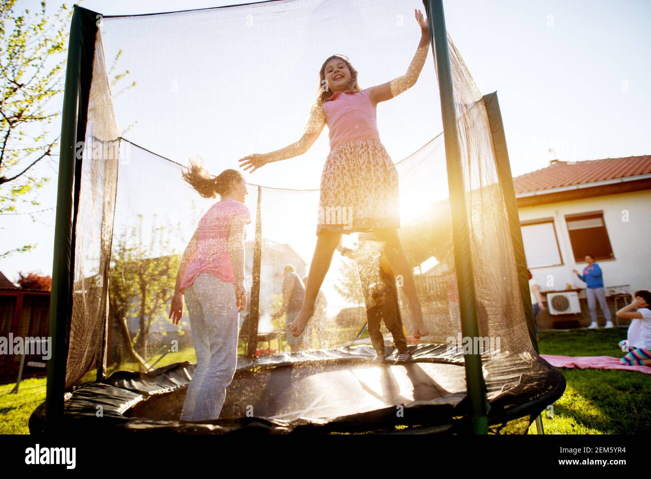 Zwei entzückende verspielte Mädchen und ein kleines Kleinkind Junge sind Mit Zeit ihres Lebens auf einem Trampolin, während die Der Rest ihrer Familie grillen und havin Stockfoto