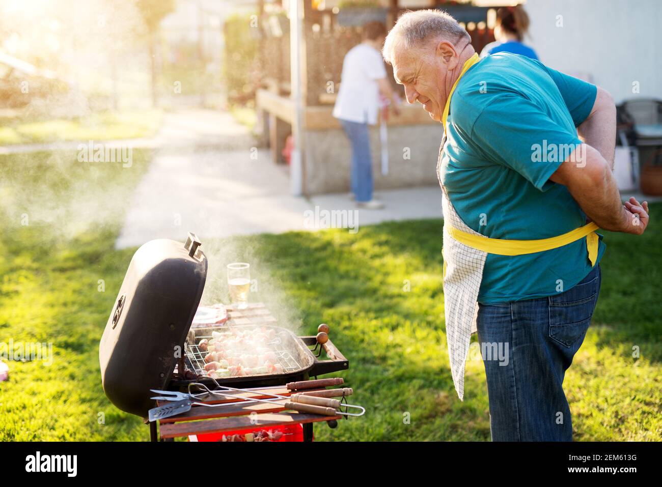 Älterer, zufriedener Mann tieing seine Schürze, während er stolz prüft, ob sein gegrilltes Fleisch fertig ist. Stockfoto