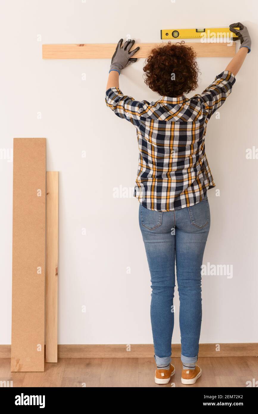 Unkenntlich Frau verwendet eine Ebene, um genau eine Holzdiele auf einer weißen Wand in einer Wohnung mit natürlichem Tageslicht zu platzieren. Stockfoto