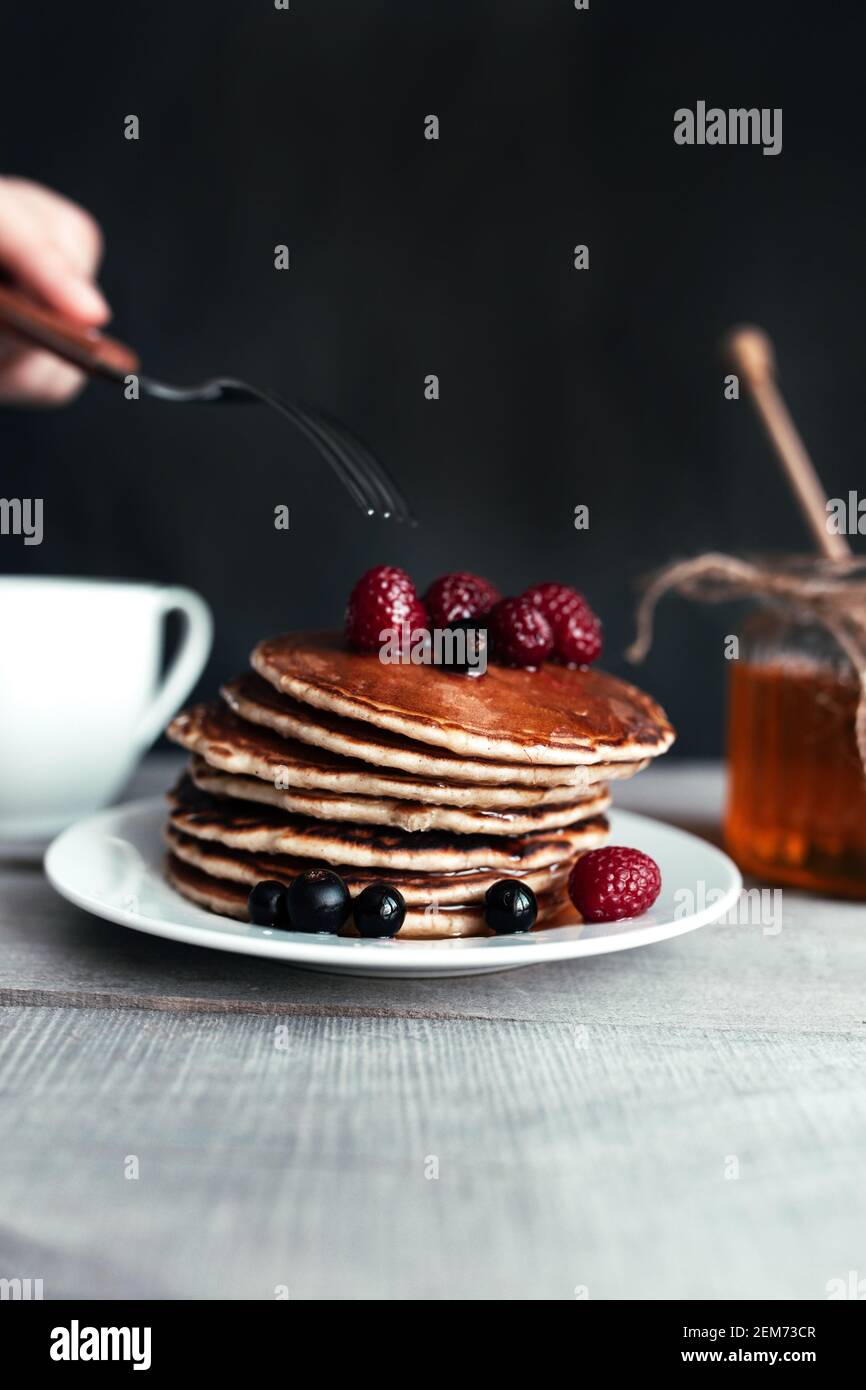 Pfannkuchen mit Beeren und Honig auf weißem Teller, Handgabel, Löffel im Glas, Holztisch, Tasse Tee. Hochwertige Fotos Stockfoto