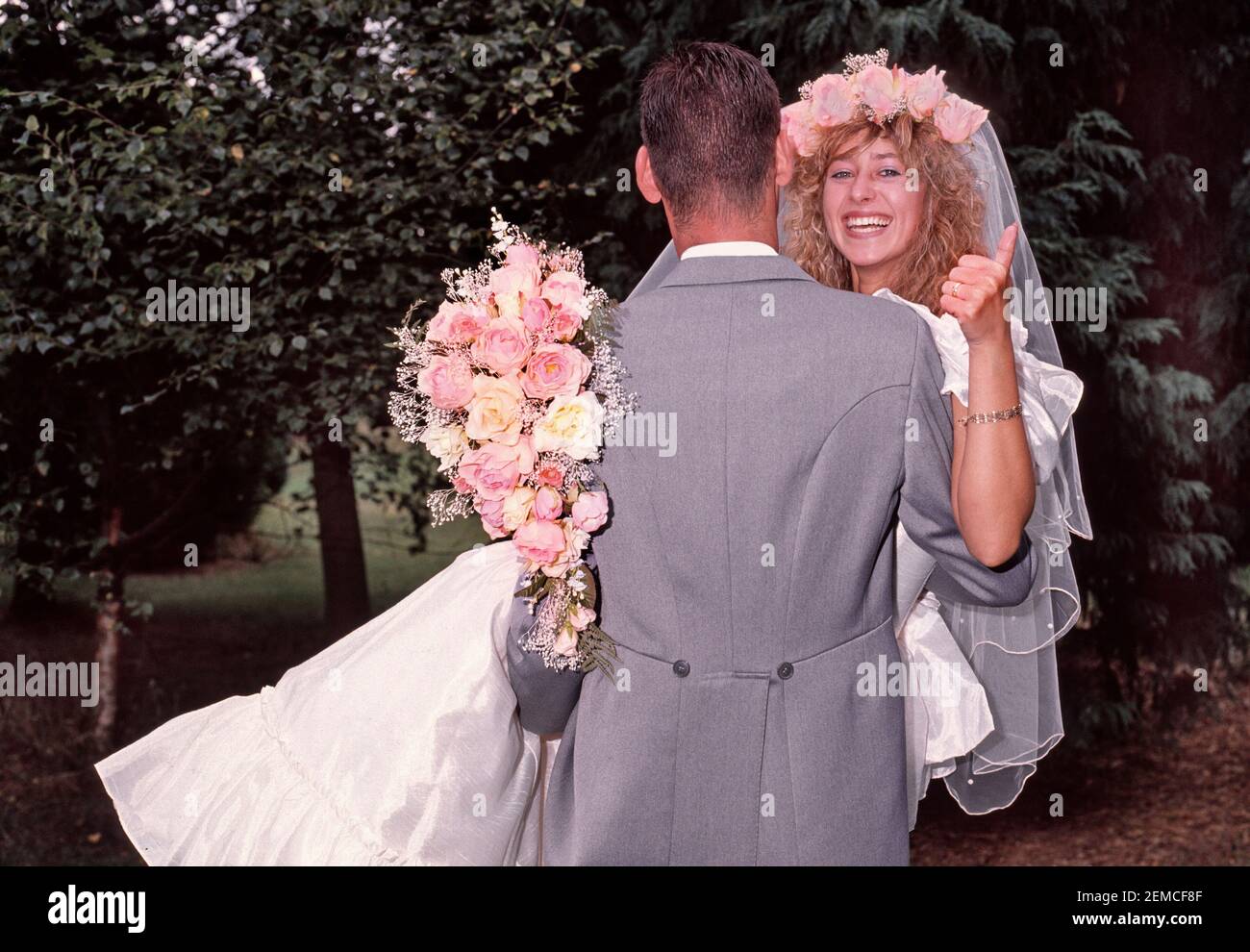 Hochzeit im Jahr 1989 von jungen Modell freigelassene Paar posiert im Freien Für Fotografien in 80s Mode Braut im Hochzeitskleid mit Blumenstrauß gibt Daumen nach oben getragen vom Bräutigam Rückansicht Grauer Morgenanzug und das Tragen einer lächelnden glücklichen Braut historischen 1980s wie wir in Essex England archivierten Stockfoto