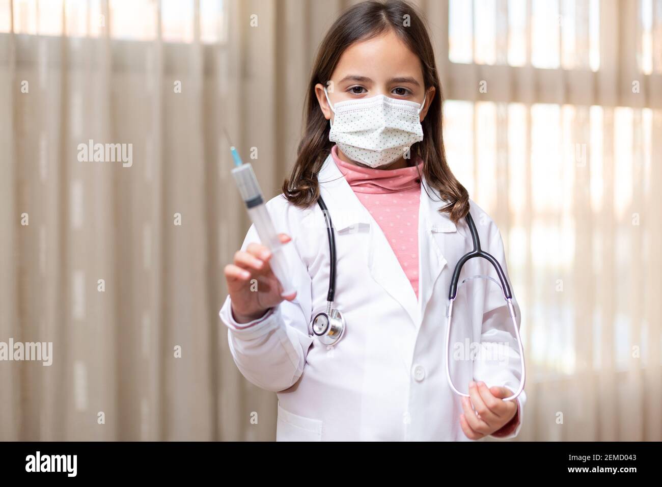 Kleines Mädchen, gekleidet als Arzt und medizinische Maske mit einer Spritze in der Hand. Konzept der Gesundheit. Leerzeichen für Text. Stockfoto
