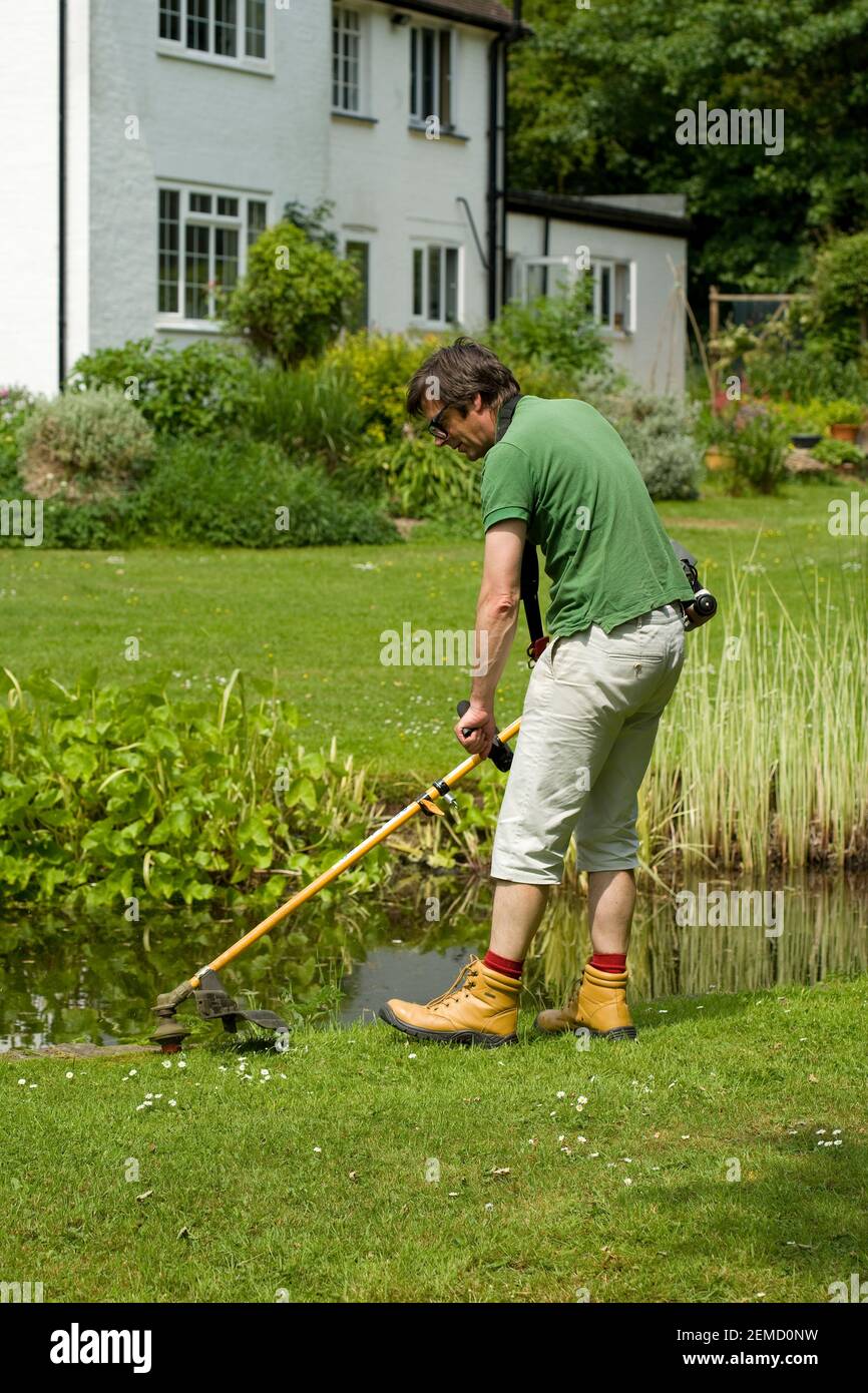 Ein Mann, der einen Fadenschneider am Teich im Garten benutzt. Auch bekannt als Strimmer oder Unkrautfresser. Stockfoto