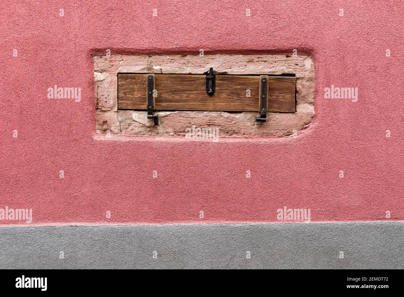 Kleines, geschlossenes und mit Holzverschalungen verkabeltes Kellerfenster in einer Steinmauer, umgeben von rosa Render, über einer grau gerenderten Sockelleiste im Elsass, Frankreich Stockfoto