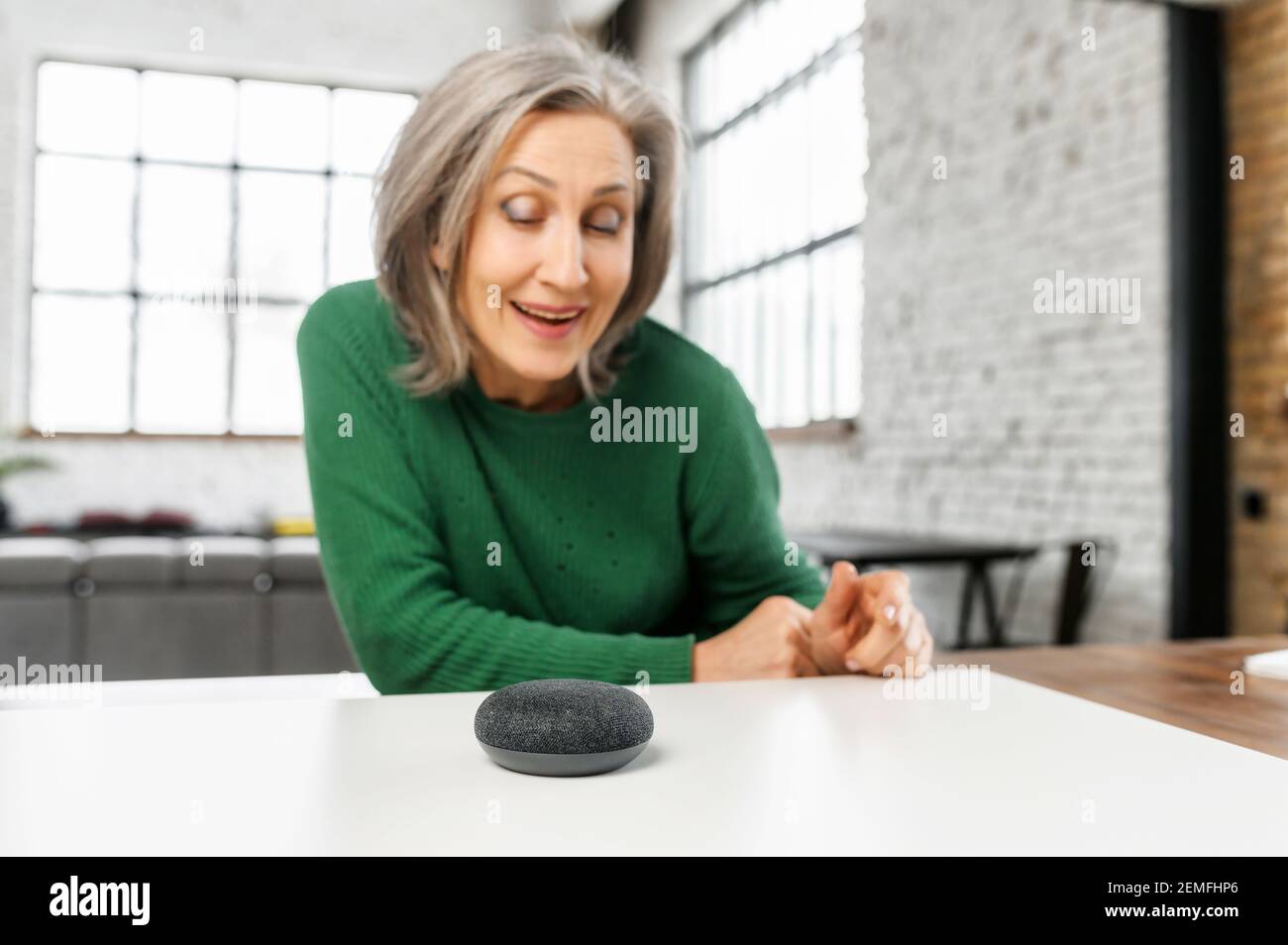 Ältere ältere moderne Frau verwendet Sprachbefehle für drahtlose intelligente Lautsprechersteuerung. Smart Home und Technologiekonzept Stockfoto