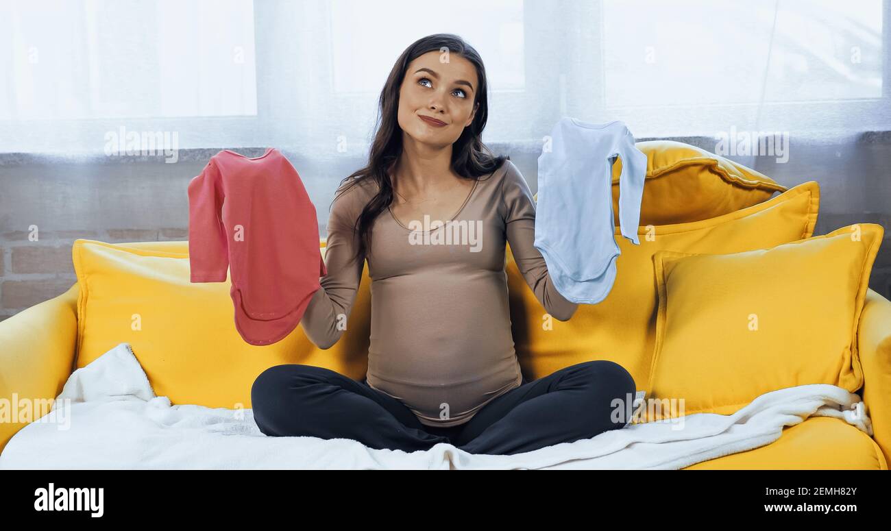 Nachdenklich schwangere Frau hält Baby-Bodysuits im Wohnzimmer Stockfoto
