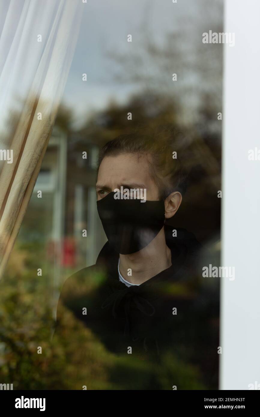 Ein junger Mann mit Gesichtsmaske, der während seiner Selbstisolation zu Hause aus dem Fenster schaut. Covid-19 Corona Virus Isolation Einsamkeit Konzept Stockfoto