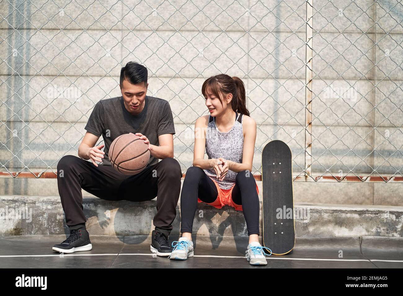 Junge asiatische Erwachsene männliche Basketballspieler und weibliche Skateboarder sitzen Auf der Gerichtsseite reden chatten Stockfoto