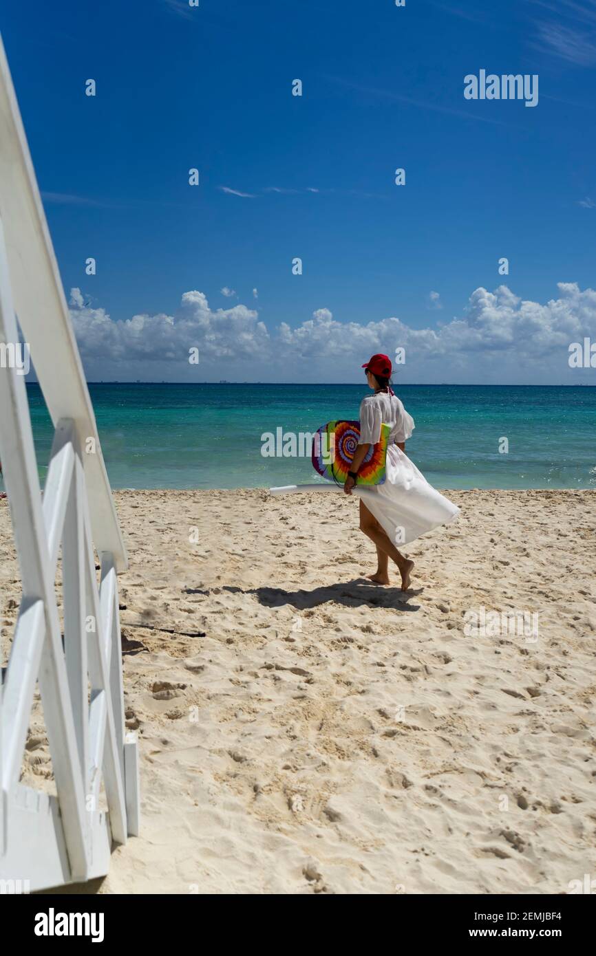 Eine attraktive Frau spaziert an einem tropischen Strand mit einem weißen Kleid und einem Surfbrett in Mexiko. Karibisches Meer und blauer Himmel im Hintergrund. Konzeptreise Stockfoto