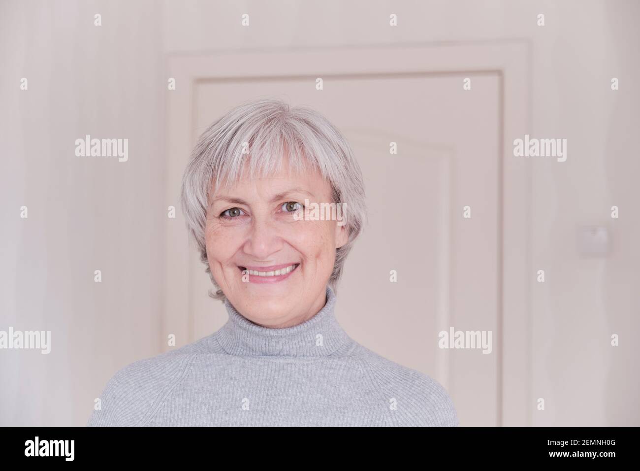 Ein Nahaufnahme Porträt einer lächelnden und Blick auf die Kamera ältere kaukasische Frau mit kurzen grauen Haaren auf einem hellen Hintergrund. Stockfoto